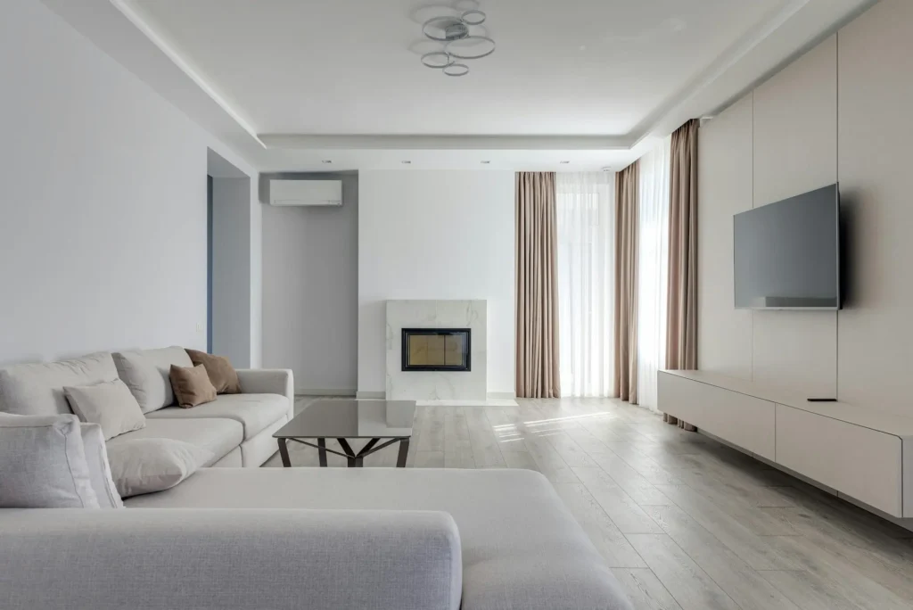 Elegante salón de tonos neutros con un sofá chaise longue gris, pared principal con televisión plana integrada, chimenea revestida de mármol y grandes ventanales vestidos, vivienda gestionada por Inmobiliaria Barcelona.