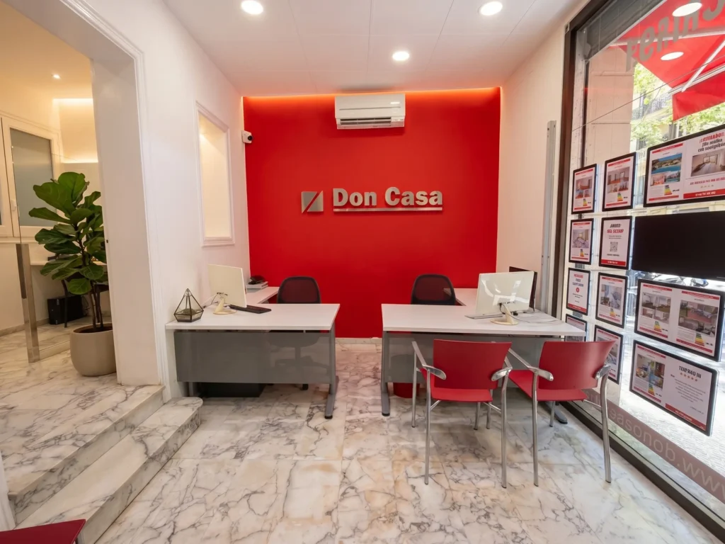 Interior de la oficina de Inmobiliaria Barcelona en Gràcia, con un diseño moderno, pared corporativa roja, puestos de trabajo ordenados y escaparate con propiedades destacadas.