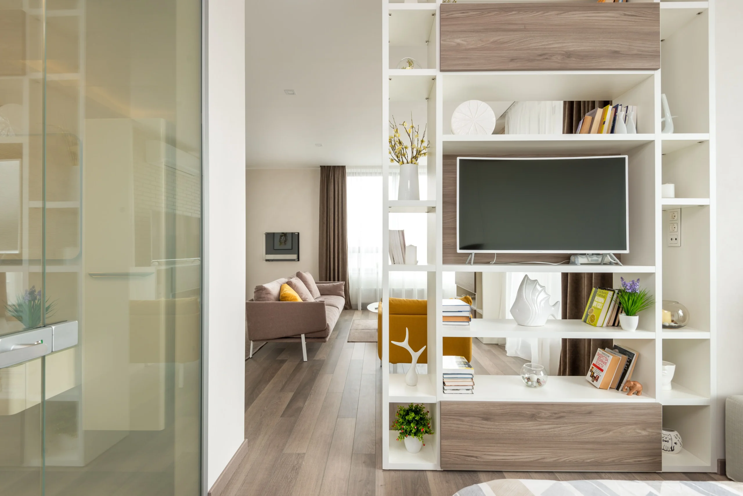 Vista interior de un piso moderno en Cornellà, Barcelona, a través de una partición de vidrio. En primer plano, una estantería blanca y de madera llena de libros, arte y un televisor. Al fondo, un salón de planta abierta con un sofá gris y un sillón amarillo. El piso cuenta con suelos de madera clara. Esta propiedad está listada por Don Casa Inmobiliaria.