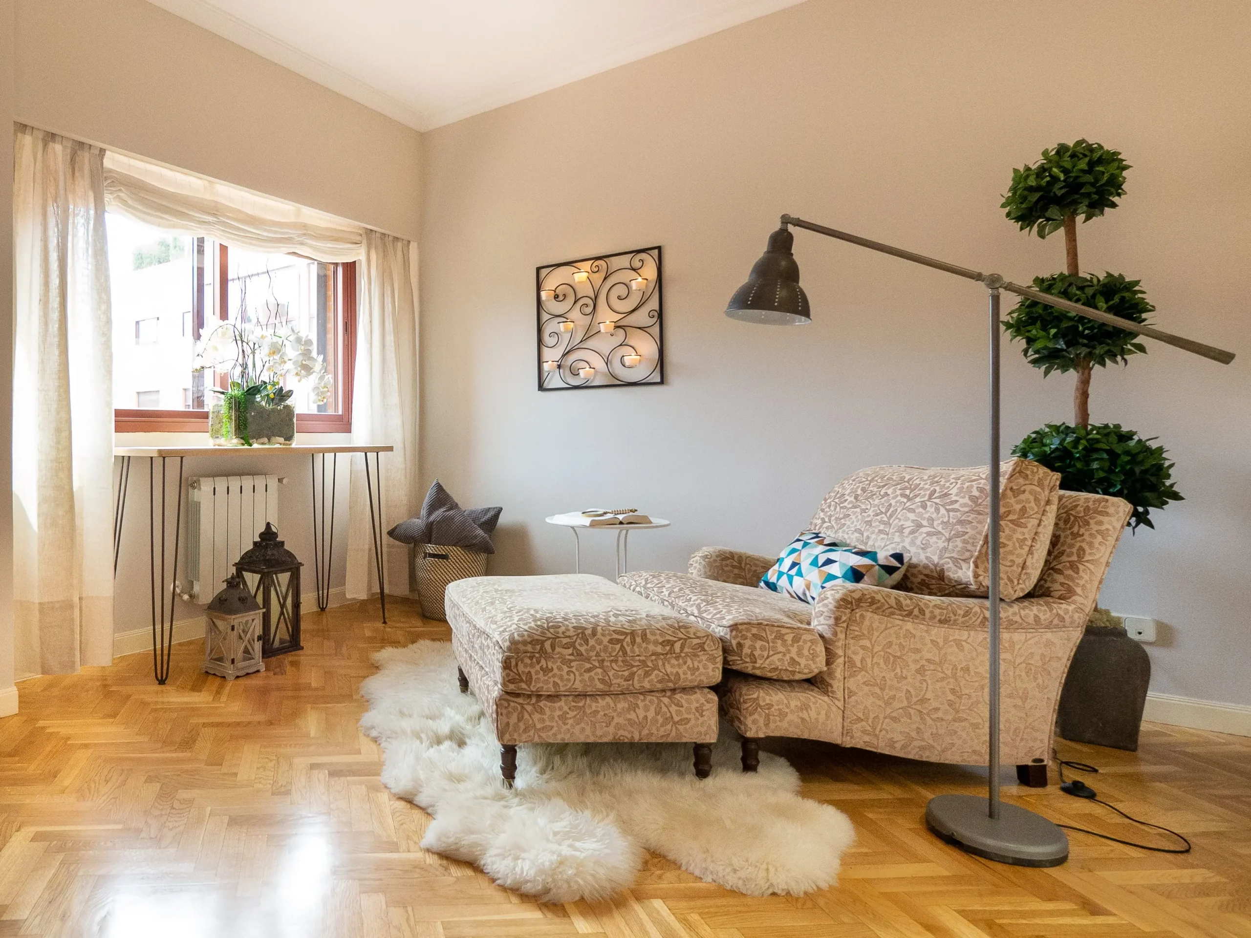 Se muestra un rincón acogedor de una sala de estar o espacio de lectura en un piso en venta en Sagrada Familia. En primer plano, se encuentran un sillón y un otomano tapizados con un patrón floral, situados sobre una suave alfombra de piel de oveja blanca, que a su vez descansa sobre un suelo de parqué de espiga. Una lámpara de pie industrial articulada de color gris se inclina sobre el sillón. En la pared, hay una decoración de metal forjado con múltiples candelabros para velas. A la izquierda, una ventana con cortinas transparentes deja pasar la luz natural, iluminando una orquídea en una mesa auxiliar y linternas decorativas en el suelo. A la derecha, un macetero con topiarios de boj. Esta imagen, presentada por Inmobiliaria Don Casa, representa el confort de esta propiedad.